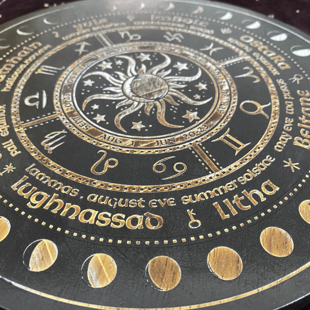 Close-up of a black and gold Wheel of the Year board highlighting the central sun and moon design, zodiac symbols, and sabbat names including Lughnasadh and Litha. Detailed pagan seasonal wheel with intricate gold engraving and moon phase markers.