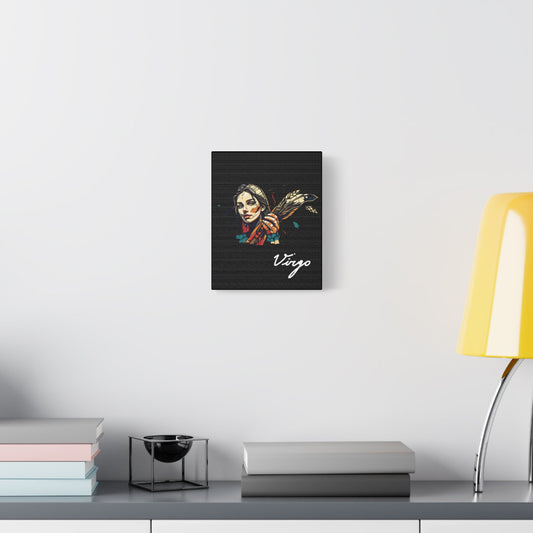 Black canvas wall art print of colorful abstract Virgo woman holding wheat sheaf, serene expression, stylish hair, and cosmic splatters on dark background, featuring "Virgo" in elegant white cursive displayed in a modern home décor setting above a console table with books and yellow lamp.