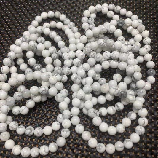 White howlite gemstone bracelets: multiple strands of polished round beads in creamy white with subtle gray-black veining and marbling, displayed in a loose pile on a dark woven mat. Natural calming crystal jewelry for stress relief, patience, and sleep support.