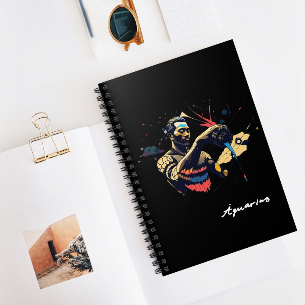 Black spiral-bound notebook featuring a vibrant Aquarius zodiac illustration of a water bearer pouring water in gold, blue, and red tones with abstract splashes, “Aquarius” written in white script at the bottom, styled on a clean desk with sunglasses and stationery items.