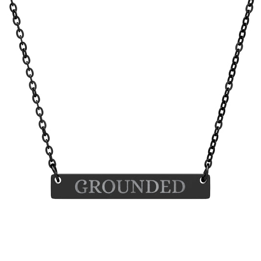 Black bar necklace with engraved uppercase text "GROUNDED" on a matte black pendant, suspended on a delicate black chain. Minimalist motivational affirmation jewelry for grounding, stability, and root chakra energy.