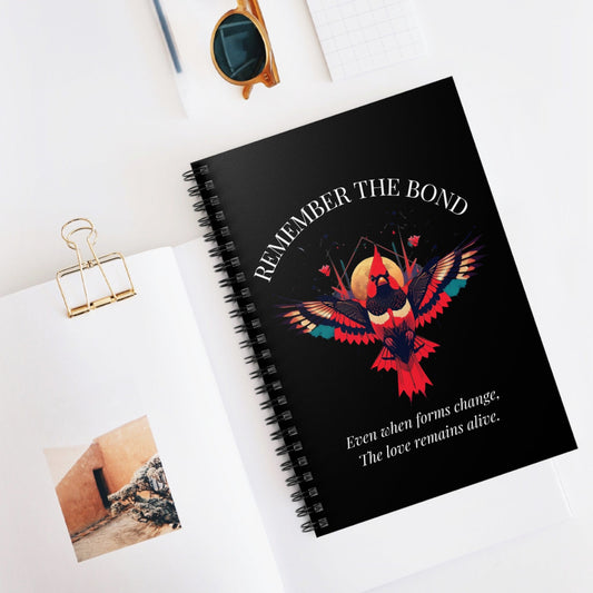 Spiral notebook with 'Remember The Bond' inspirational bird cover design and quote 'Even when forms change, The love remains alive' styled on a white desk with accessories.