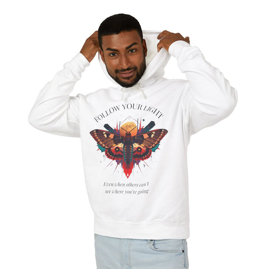 Young man with short hair pulling up the hood of a white hoodie featuring vibrant multicolored moth/butterfly graphic, geometric sun/moon elements, "FOLLOW YOUR LIGHT" arched text above, motivational quote "Even when others can't see where you're going" below
