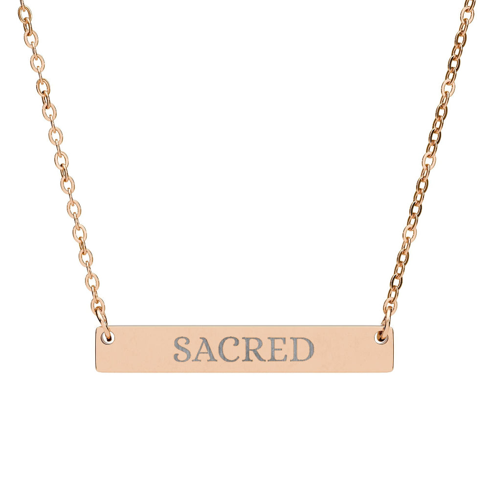 Rose gold bar necklace with engraved uppercase text "SACRED" on a sleek matte pendant, suspended on a delicate matching rose gold chain. Minimalist spiritual affirmation jewelry for sacred energy and divine connection.
