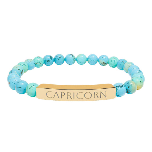 Bracelet with turquoise beaded stretch strand in soft aqua-blue tones with natural veining and speckles, featuring a gold-plated curved bar engraved "CAPRICORN" in uppercase letters. Zodiac-inspired healing crystal jewelry for ambition, discipline, and grounding.