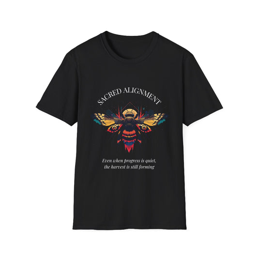 Black unisex cotton t-shirt laid out flat featuring bold "Sacred Alignment" graphic: vibrant abstract bee with detailed wings and body, colorful geometric accents, and dynamic paint splatters in red, orange, teal, blue, and gold tones bursting outward around an orange moon background. Arched white text "SACRED ALIGNMENT" above and smaller inspirational quote "Even when progress is quiet, the harvest is still forming" below. Motivational spiritual empowerment crewneck tee.
