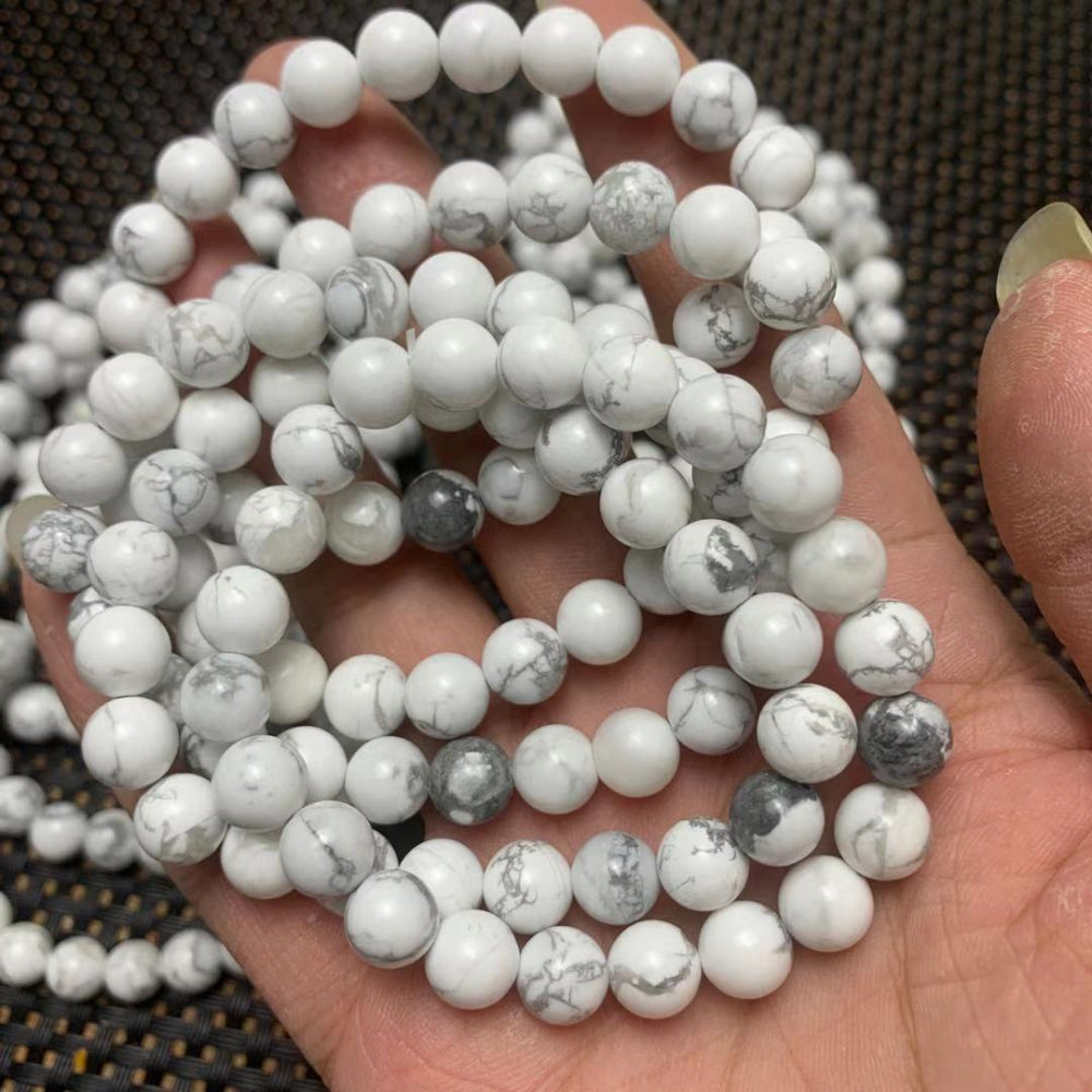 Close-up of white howlite round gemstone bead bracelets with natural gray veining, polished beads displayed in hand on a dark textured background.