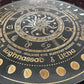 Close-up of a black and gold Wheel of the Year board highlighting the central sun and moon design, zodiac symbols, and sabbat names including Lughnasadh and Litha. Detailed pagan seasonal wheel with intricate gold engraving and moon phase markers.