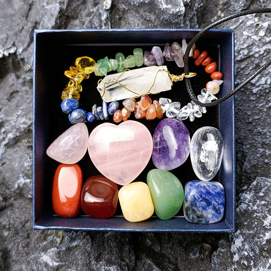 Chakra healing crystal gift set in navy blue gift box on natural rock surface – rose quartz heart, tumbled gemstones (jasper, carnelian, citrine, aventurine, lapis, amethyst, quartz), gold-wrapped clear quartz pendant on black cord, chakra chip bracelet, premium spiritual wellness collection
