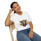 White unisex cotton t-shirt modeled by smiling woman with braided hair, seated pose, hand on cheek, featuring bold Leo zodiac graphic: vibrant abstract lion head portrait with powerful flowing mane, fierce intense eyes, regal expression, geometric accents, and dynamic multicolored paint splatters in gold, red, navy, teal, and black tones exploding outward. Elegant black cursive "Leo" text below. Astrology zodiac motivational crewneck tee.