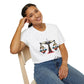 White unisex cotton t-shirt modeled by smiling woman with braided hair, seated pose, hand on cheek, featuring bold Libra zodiac graphic on chest: vibrant abstract scales of justice with balanced arms, intricate geometric and triangular motifs, central axis design, symmetrical layout, and dynamic multicolored paint splatters in gold, red, navy, teal, and black tones bursting outward. Elegant black cursive "Libra" text below. Astrology zodiac motivational crewneck tee.