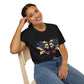 Black unisex cotton t-shirt modeled by smiling woman with braided hair, seated pose, hand on cheek, featuring bold Gemini zodiac graphic: vibrant abstract twin female faces with flowing hair, intense expressions, geometric patterns, and dynamic multicolored paint splatters in gold, red, navy, teal, and black tones exploding outward to form butterfly wings. Elegant white cursive "Gemini" text below. Astrology zodiac motivational crewneck tee.