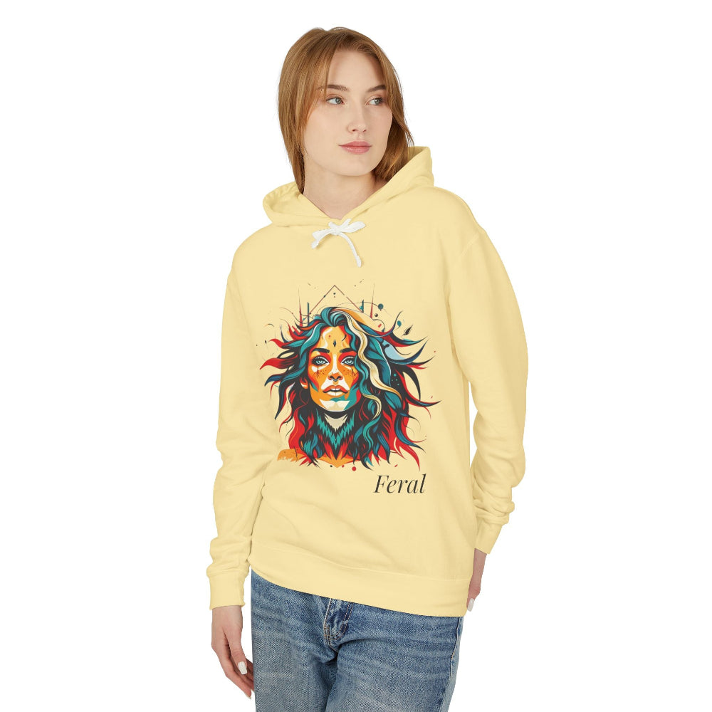 Young woman with long hair modeling yellow hoodie featuring vibrant artistic portrait of a fierce woman with curly multicolored flowing hair, teal red blue orange face accents, large hoop earrings, third-eye symbol, "Feral" text below