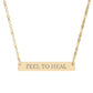 Gold bar necklace with engraved uppercase text "FEEL TO HEAL" on a sleek matte gold pendant, suspended on a delicate matching gold chain. Minimalist motivational affirmation jewelry for emotional healing and self-care.