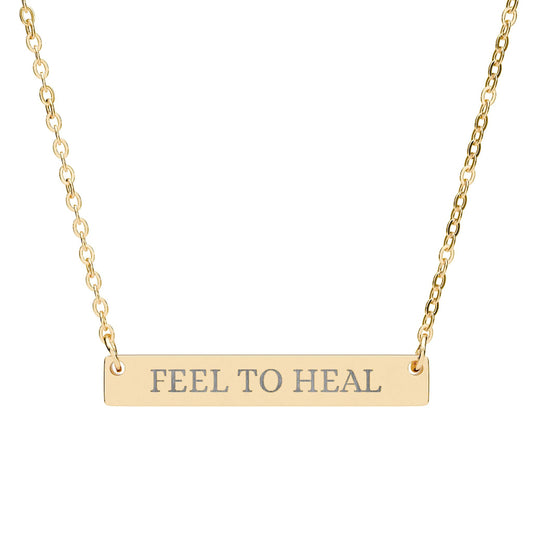 Gold bar necklace with engraved uppercase text "FEEL TO HEAL" on a sleek matte gold pendant, suspended on a delicate matching gold chain. Minimalist motivational affirmation jewelry for emotional healing and self-care.