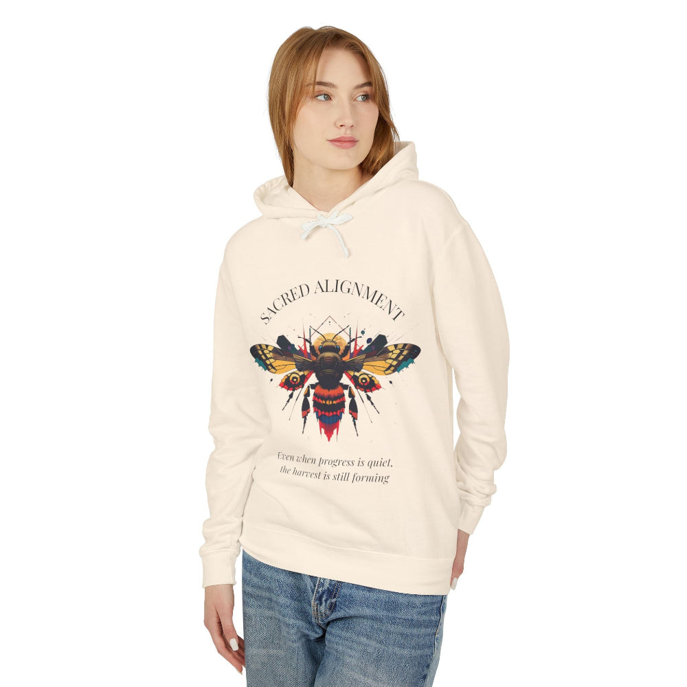 Woman wearing an off-white hoodie featuring a vibrant bee graphic with the text "SACRED ALIGNMENT" above and the quote "Even when progress is quiet, the harvest is still forming" below. Casual streetwear style with blue jeans.