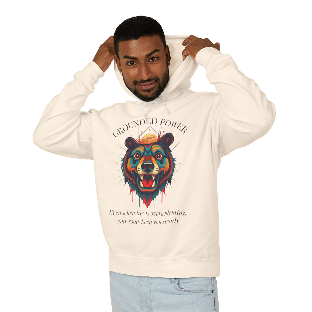 Young man with short hair pulling up the hood of a cream hoodie featuring vibrant multicolored bear head graphic, roaring expression, fur in blue orange teal splashes, orange moon above, "GROUNDED POWER" arched text, motivational quote about roots and steadiness below