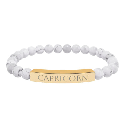Bracelet with polished white howlite beads featuring natural gray-black veining and marbling, paired with a gold-plated curved bar engraved "CAPRICORN" in uppercase letters. Stretch-fit zodiac-inspired crystal jewelry.
