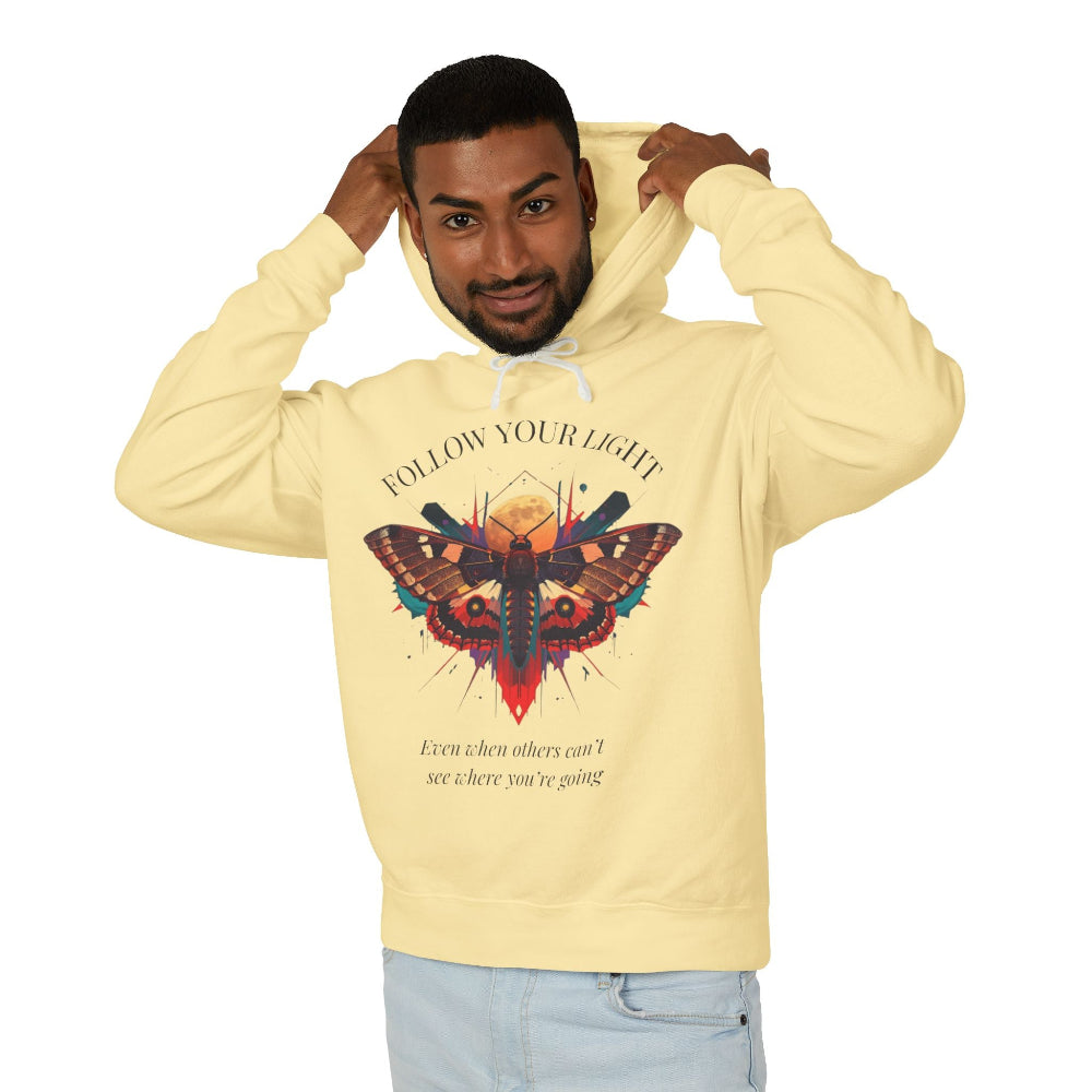 Young man with short hair pulling up the hood of a pale yellow hoodie featuring vibrant multicolored moth/butterfly graphic, geometric sun/moon elements, "FOLLOW YOUR LIGHT" arched text above, motivational quote "Even when others can't see where you're going" below