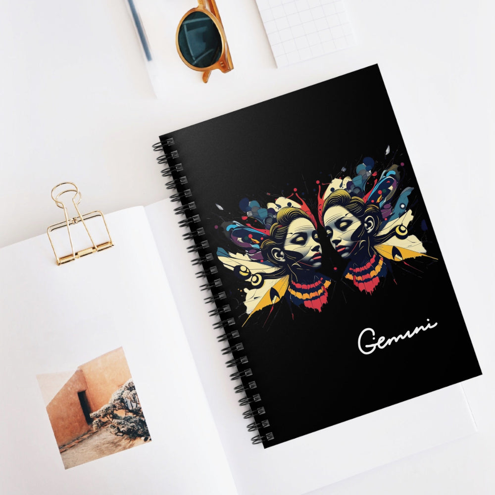 Notebook with a colorful abstract Gemini twin faces design, flowing hair, cosmic accents, and paint splatters on a black background, featuring the word "Gemini" in elegant white cursive, placed on a minimal white desk with sunglasses and stationery items.
