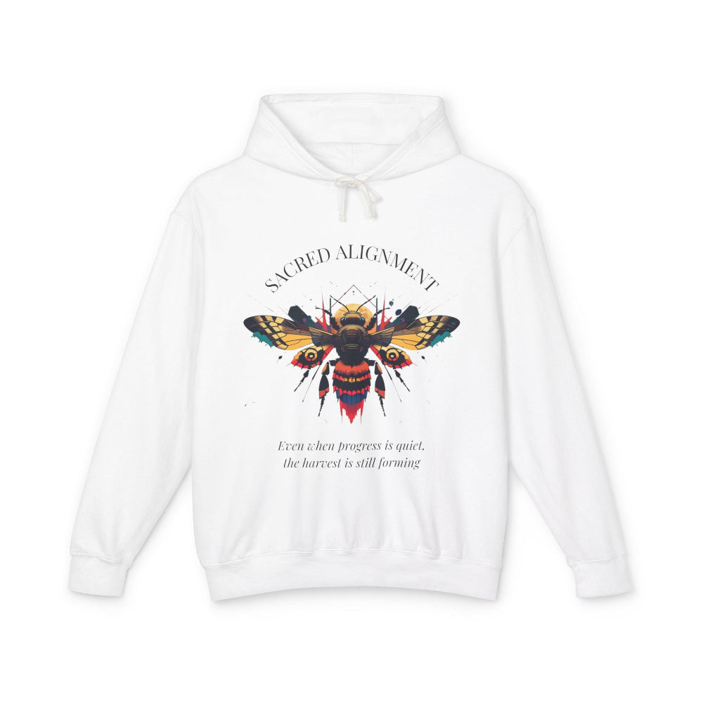 White hoodie laid flat, featuring a vibrant colorful bee graphic with the text "SACRED ALIGNMENT" arched above and the quote "Even when progress is quiet, the harvest is still forming" below. Motivational streetwear apparel.