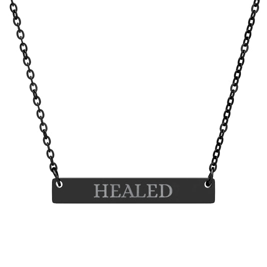 Black bar necklace with engraved uppercase text "HEALED" on a matte black pendant, suspended on a delicate black chain. Minimalist motivational affirmation jewelry for healing, recovery, and emotional restoration.