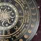 Close-up detail of a black and gold Wheel of the Year board showing the sun and moon illustration, zodiac symbols, sabbat names including Ostara, Beltane, and Litha, along with moon phase markers. Pagan seasonal wheel with intricate gold engraving.