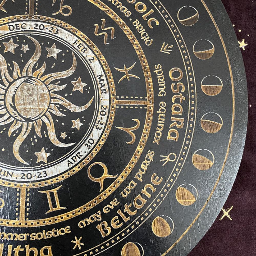 Close-up detail of a black and gold Wheel of the Year board showing the sun and moon illustration, zodiac symbols, sabbat names including Ostara, Beltane, and Litha, along with moon phase markers. Pagan seasonal wheel with intricate gold engraving.
