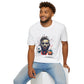 White unisex cotton t-shirt modeled by smiling man with curly hair and beard, hand in pocket, featuring bold "Primal" graphic on chest: stylized intense male portrait with stylish hair, vibrant multicolored geometric face in red, orange, teal, blue and gold tones, dynamic paint splatters and abstract shards exploding outward. Elegant white cursive "Primal" text below. Primal masculine energy motivational crewneck tee.