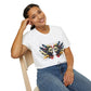 White unisex cotton t-shirt modeled by smiling woman with braided hair, seated pose, hand on cheek, featuring bold Gemini zodiac graphic: vibrant abstract twin female faces with flowing hair, intense expressions, geometric patterns, and dynamic multicolored paint splatters in gold, red, navy, teal, and black tones exploding outward. Elegant black cursive "Gemini" text below. Astrology zodiac motivational crewneck tee.