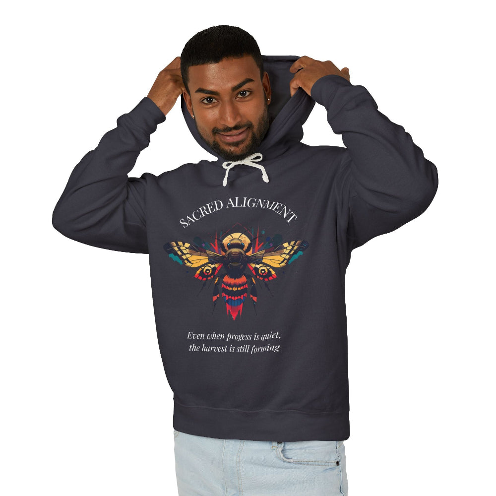 Man wearing a dark grey hoodie featuring a vibrant bee graphic with the text "SACRED ALIGNMENT" above and the quote "Even when progress is quiet, the harvest is still forming" below. Casual streetwear style with light jeans.
