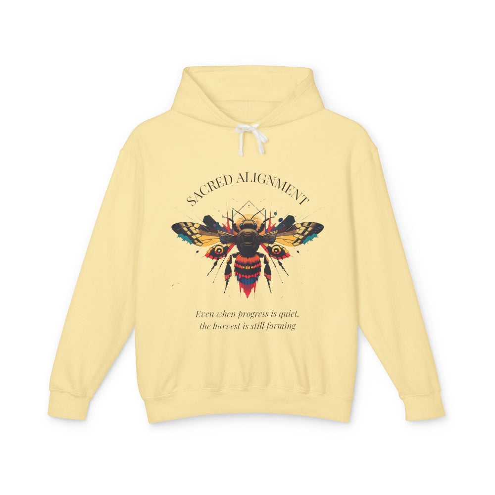 Yellow hoodie laid flat, featuring a vibrant colorful bee graphic with the text "SACRED ALIGNMENT" arched above and the quote "Even when progress is quiet, the harvest is still forming" below. Motivational streetwear apparel.