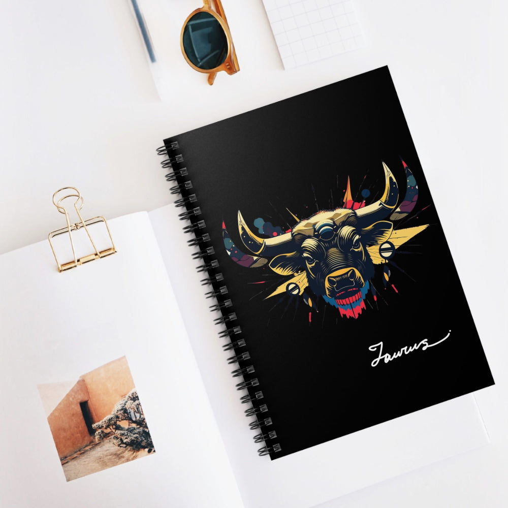 Black spiral-bound notebook with a bold Taurus zodiac bull illustration in gold, red, and blue on the cover, featuring stylized horns and artistic splashes, with “Taurus” written in white script at the bottom, placed on a clean desk with sunglasses and stationery items.