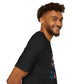 Black unisex cotton t-shirt modeled by smiling man with curly hair and beard, side profile view: bold "Primal" graphic on chest showing stylized intense male portrait with stylish hair, vibrant multicolored geometric face in red, orange, teal, blue and gold tones, dynamic paint splatters and abstract shards exploding outward. Elegant white cursive "Primal" text below. Primal masculine energy motivational crewneck tee.