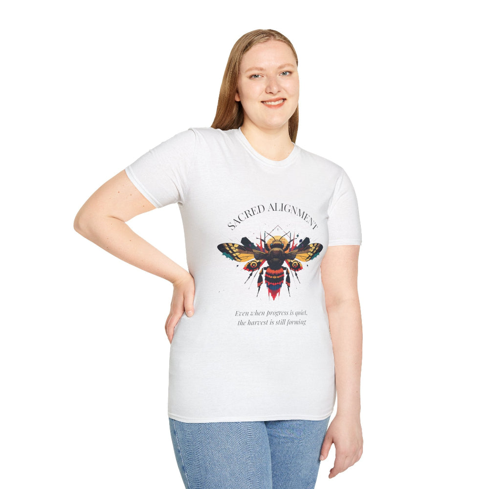 White plus-size unisex cotton t-shirt modeled by smiling woman with long blonde hair, hand on hip, featuring bold "Sacred Alignment" graphic on chest: vibrant abstract bee with detailed wings and body, colorful geometric accents, and dynamic paint splatters in red, orange, teal, blue, and gold tones around an orange moon. Arched white text "SACRED ALIGNMENT" above and smaller quote "Even when progress is quiet, the harvest is still forming" below. Motivational spiritual empowerment crewneck tee.