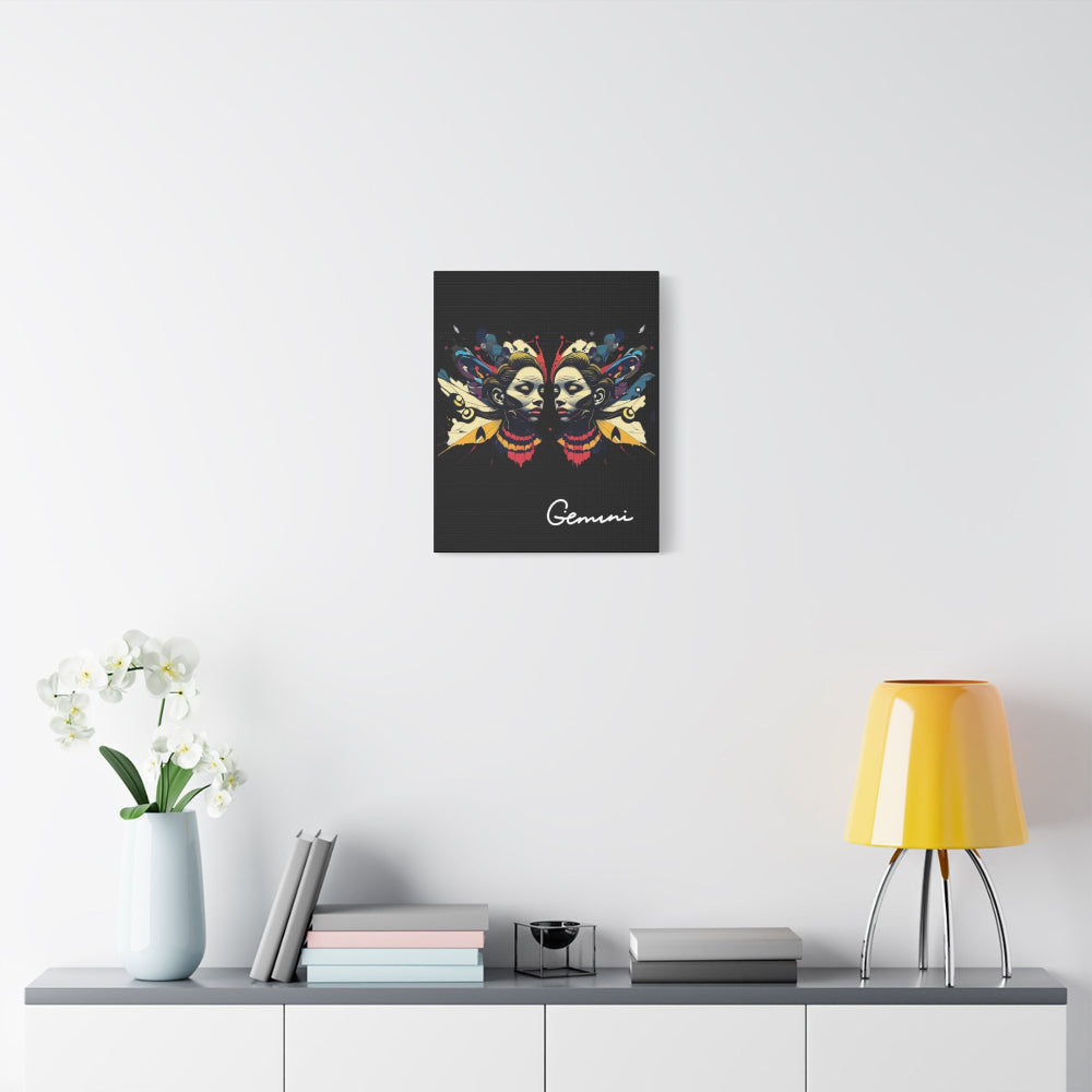 Black canvas wall art print of colorful abstract Gemini twin faces with butterfly wings and cosmic splatters on dark background, featuring "Gemini" in elegant white cursive  displayed in a modern home décor setting above a console table with books and yellow lamp.