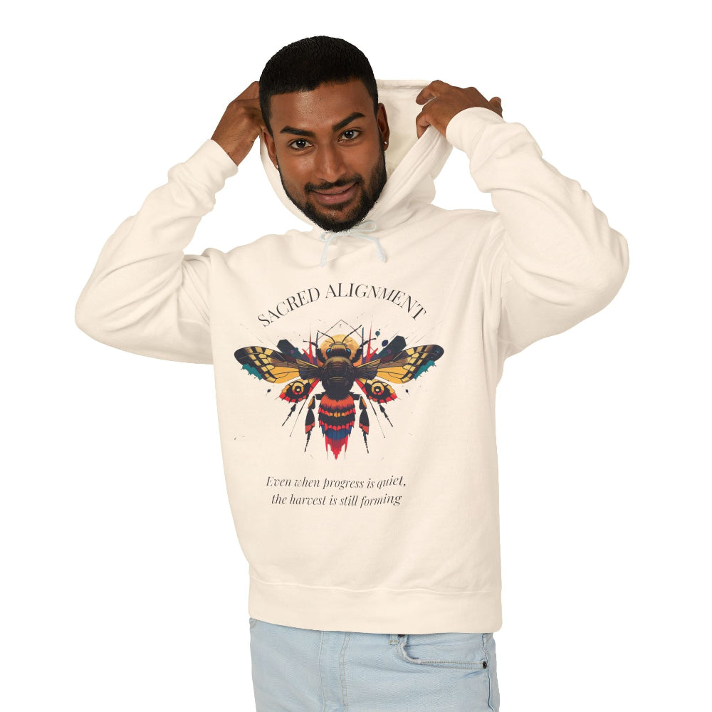 Man wearing a off-white hoodie featuring a vibrant bee graphic with the text "SACRED ALIGNMENT" above and the quote "Even when progress is quiet, the harvest is still forming" below. Casual streetwear style with light jeans.