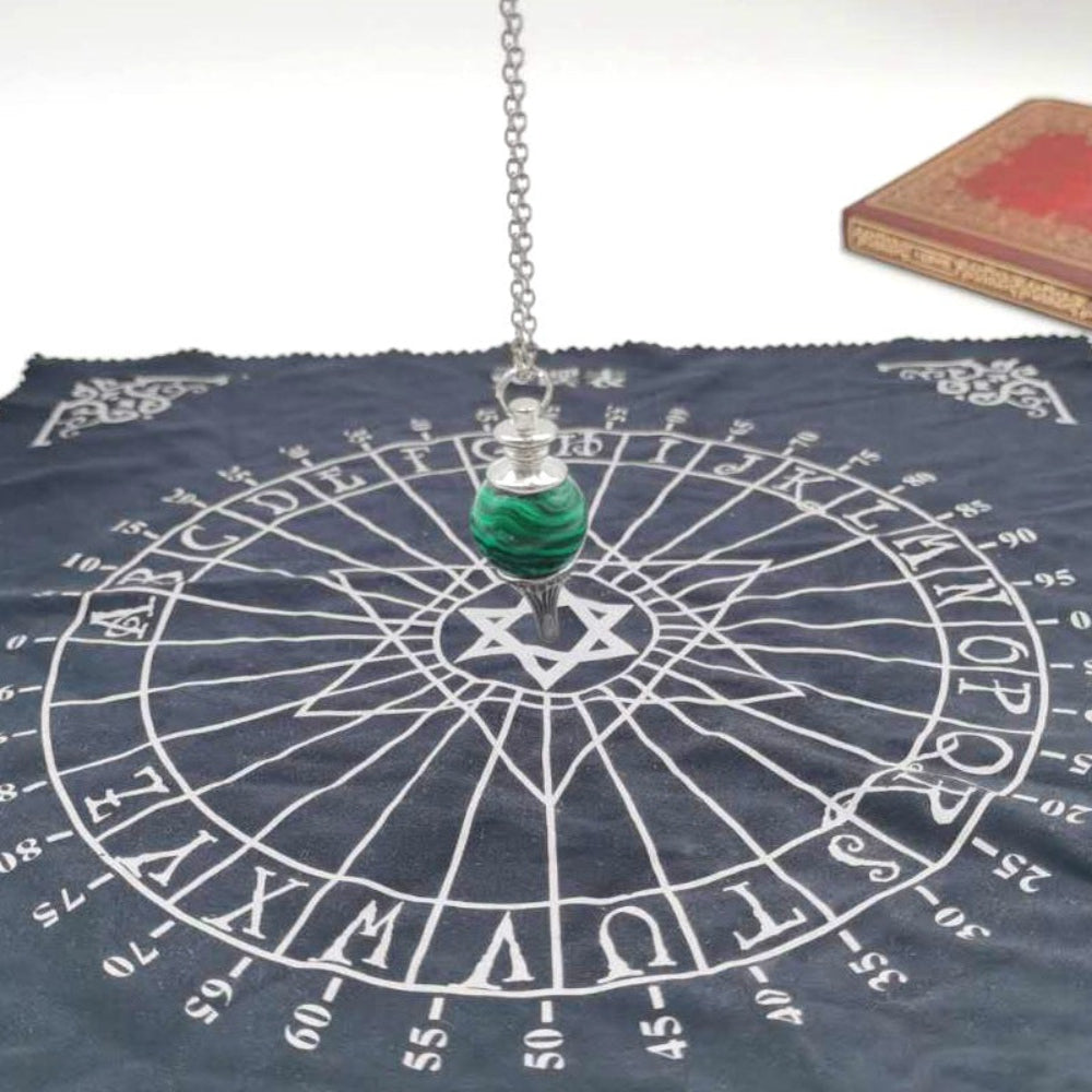 Silver pendulum with a polished green malachite sphere hanging over the center of a black spirit board cloth. The board features letters A-Z, numbers 0-9, "YES" and "NO", and a central Star of David symbol. Divination and dowsing tool setup.