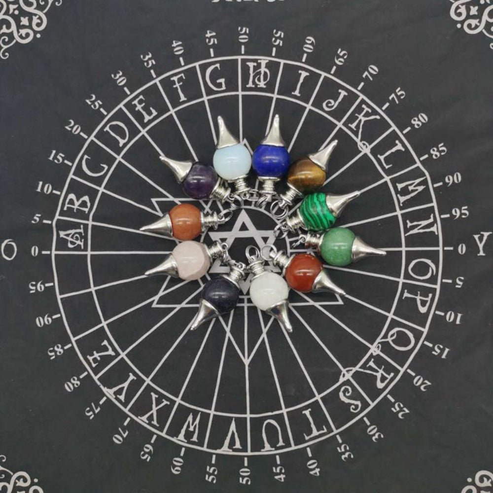 Collection of silver pendulums with various crystal spheres (rose quartz, amethyst, lapis lazuli, opalite, sodalite, tiger's eye, malachite, green aventurine, carnelian, and more) arranged in a circle on a black spirit board chart with letters, numbers, and a central Star of David. Divination and dowsing tools.