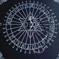 Black spirit board chart (灵摆表) with a dark blue stone pendulum resting in the center. The circular wheel features letters A-Z, numbers 0-9, "YES" and "NO", and a central Star of David symbol, framed with ornate white corner decorations. Divination and dowsing tool.