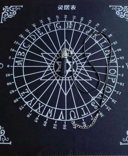 Black spirit board chart (灵摆表) with a dark blue stone pendulum resting in the center. The circular wheel features letters A-Z, numbers 0-9, "YES" and "NO", and a central Star of David symbol, framed with ornate white corner decorations. Divination and dowsing tool.