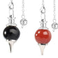 Two silver pendulums side by side: one with a glossy black onyx sphere and one with a glossy red carnelian sphere. Both have pointed metal tips and ornate silver chains with decorative acorn charms. Divination and dowsing tools.