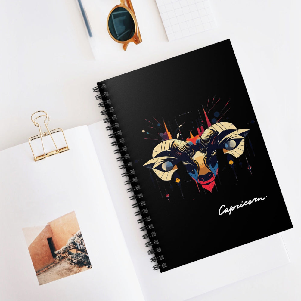 Black spiral-bound notebook featuring a bold Capricorn zodiac goat illustration in gold, blue, and red with stylized curved horns and abstract splashes, “Capricorn” written in white script at the bottom, displayed on a minimal desk with sunglasses and stationery items.