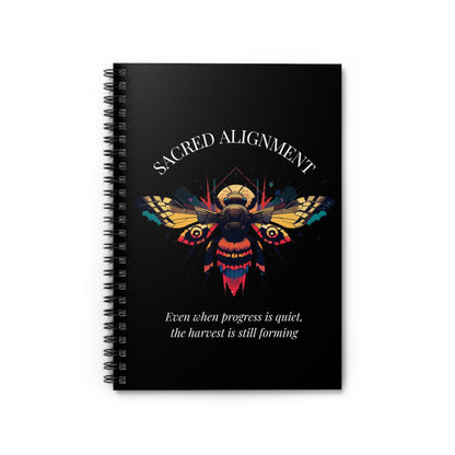 Close up of notebook with a colorful abstract sacred bee design, geometric accents, and paint splatters on a black background, featuring the words "SACRED ALIGNMENT" and quote "Even when progress is quiet, the harvest is still forming".