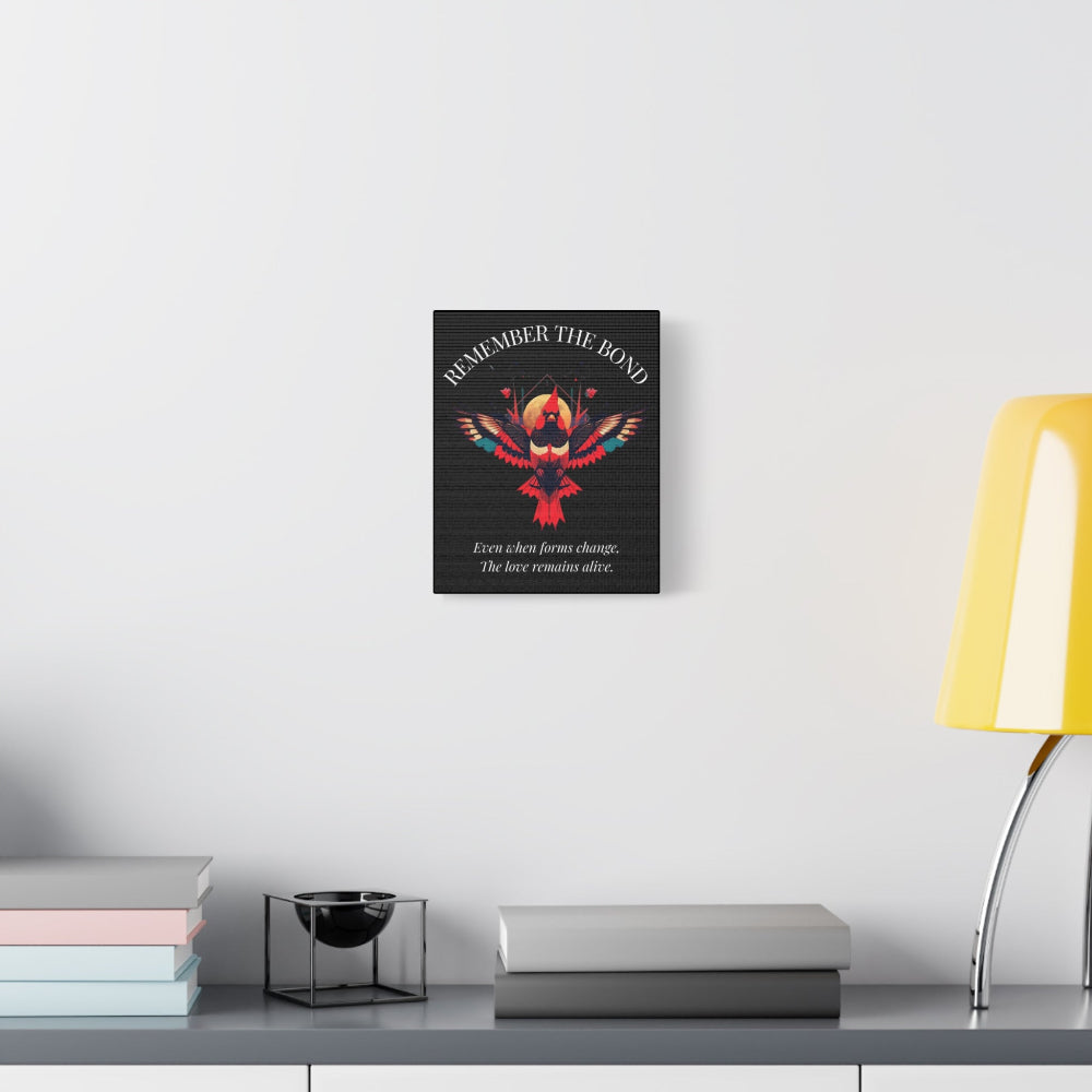 Minimalist interior scene featuring a framed wall art print with a colorful geometric phoenix and the quote "Remember the Bond – Even when forms change, the love remains alive." Styled on a light wall above a shelf with books and a yellow lamp.