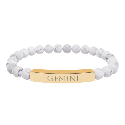 Bracelet with polished white howlite beads featuring natural gray-black veining and marbling, paired with a gold-plated curved bar engraved "GEMINI" in uppercase letters. Stretch-fit zodiac-inspired crystal jewelry.