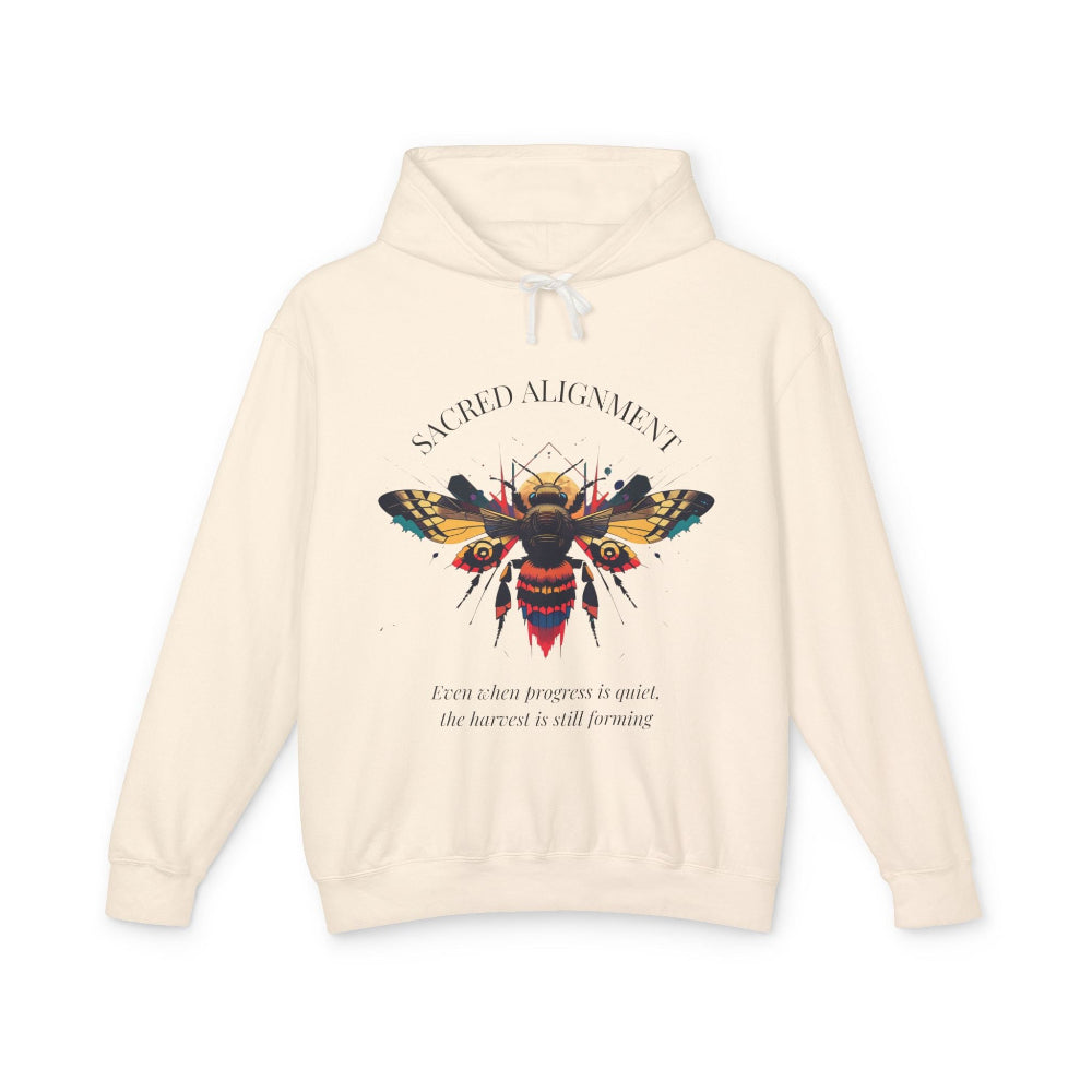 Off-white hoodie laid flat, featuring a vibrant colorful bee graphic with the text "SACRED ALIGNMENT" arched above and the quote "Even when progress is quiet, the harvest is still forming" below. Motivational streetwear apparel.
