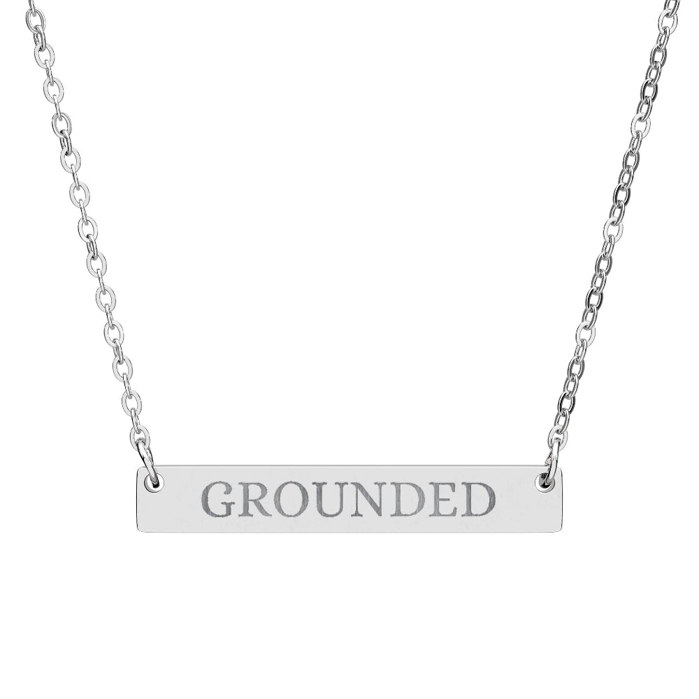 Silver bar necklace with engraved uppercase text "GROUNDED" on a sleek matte silver pendant, suspended on a delicate matching silver chain. Minimalist motivational affirmation jewelry for grounding, stability, and root chakra energy.