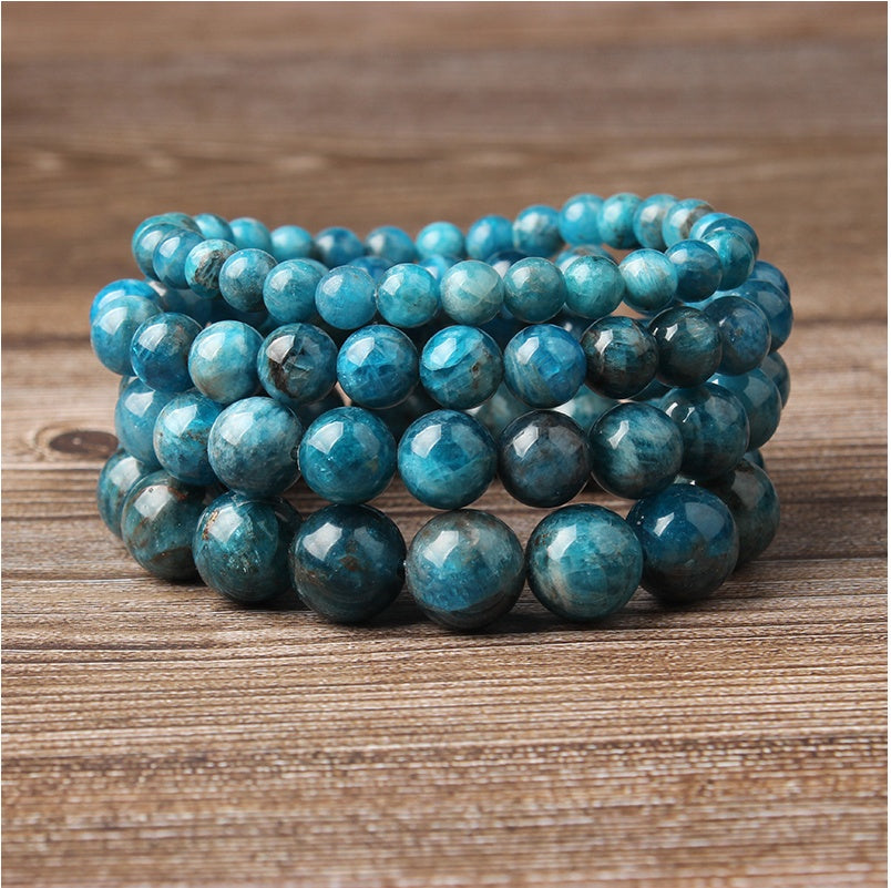 Stack of blue apatite gemstone bracelets: vibrant turquoise-blue faceted round beads with natural inclusions and subtle chatoyancy, polished shiny surfaces, stacked in multiple layers on a wooden background. Natural healing crystal jewelry for throat chakra, communication, and emotional clarity.