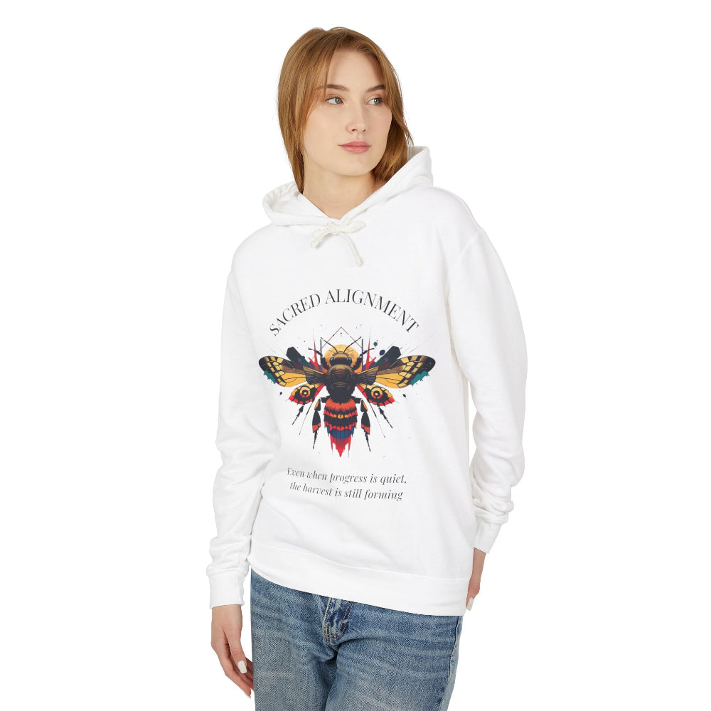 Woman wearing a white hoodie featuring a vibrant bee graphic with the text "SACRED ALIGNMENT" above and the quote "Even when progress is quiet, the harvest is still forming" below. Casual streetwear style with blue jeans.